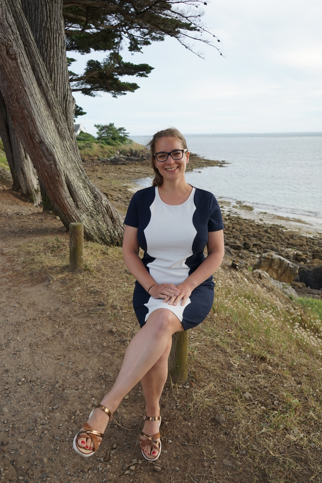 Woman sitting in a white and navy blue dress, smiling, with a background of trees, rocks and sea