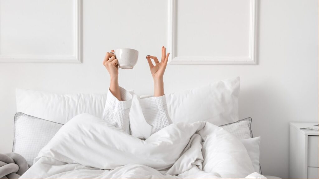 woman in a white bed, can only see arms raised, making the sign that she is fine and holding a cup